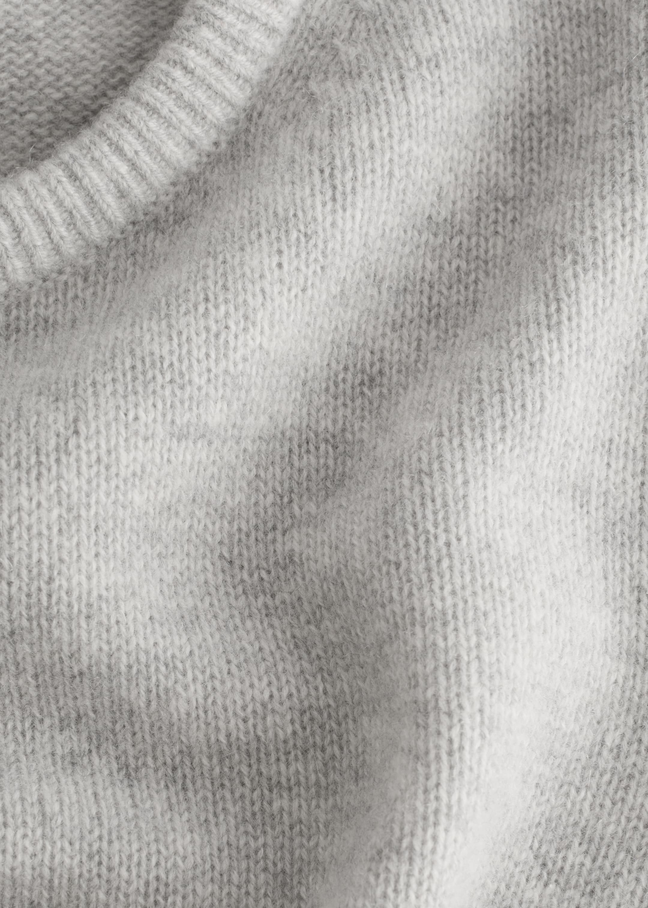 Grösseres Bild ansehen: Eine Nahaufnahme eines hellgrauen gestrickten T-Shirts zeigt ein geripptes Bündchen und einen strukturierten Körper. Der Stoff weist ein sichtbares Strickmuster und eine leicht flauschige Oberfläche auf.