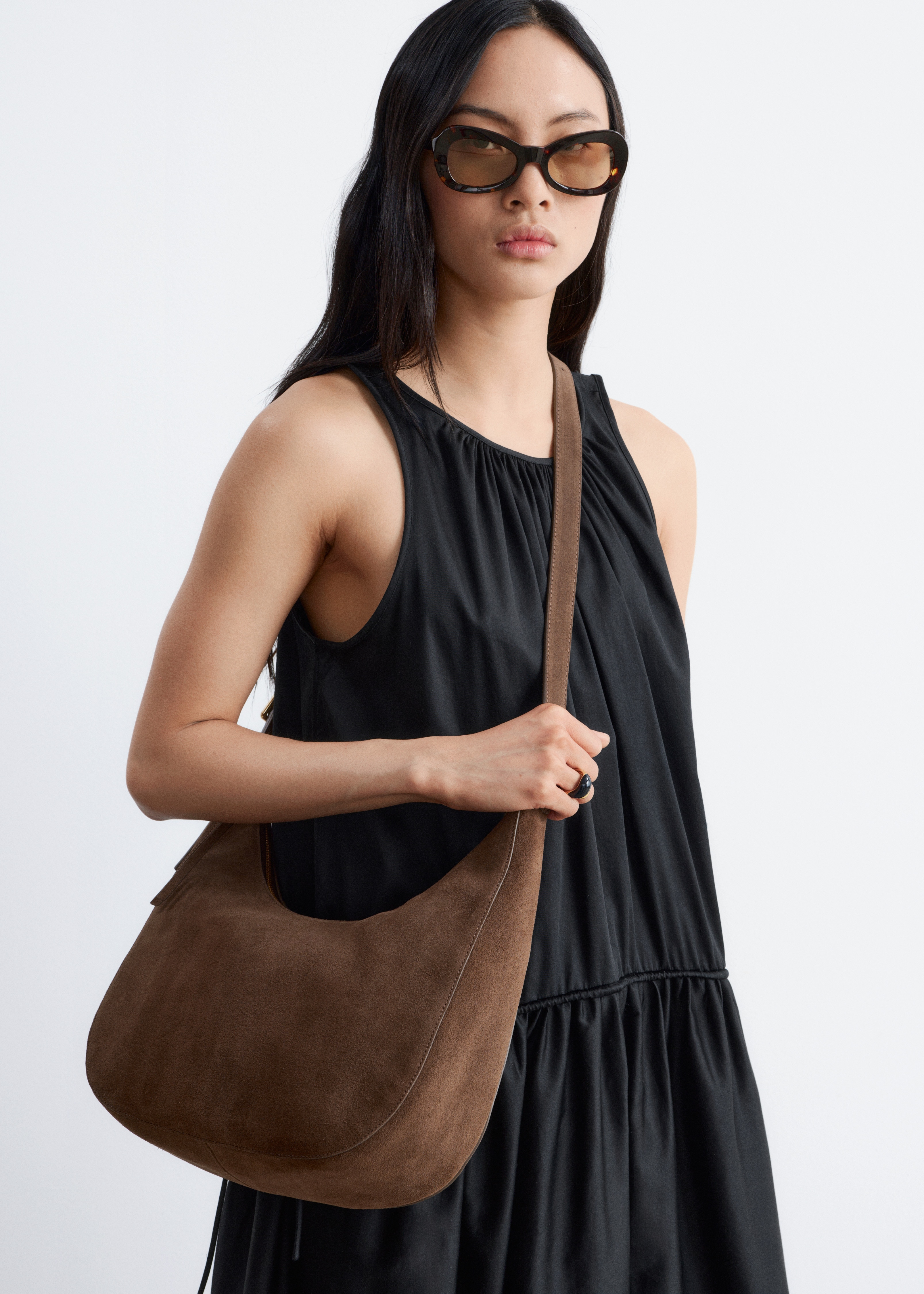 Un sac bandoulière en daim brun est suspendu, face avant, avec une forme de croissant souple. Il est doté d'une longue sangle fine et ajustable avec une boucle dorée et d'une fermeture éclair supérieure dissimulée avec un tirette à pompon brun.