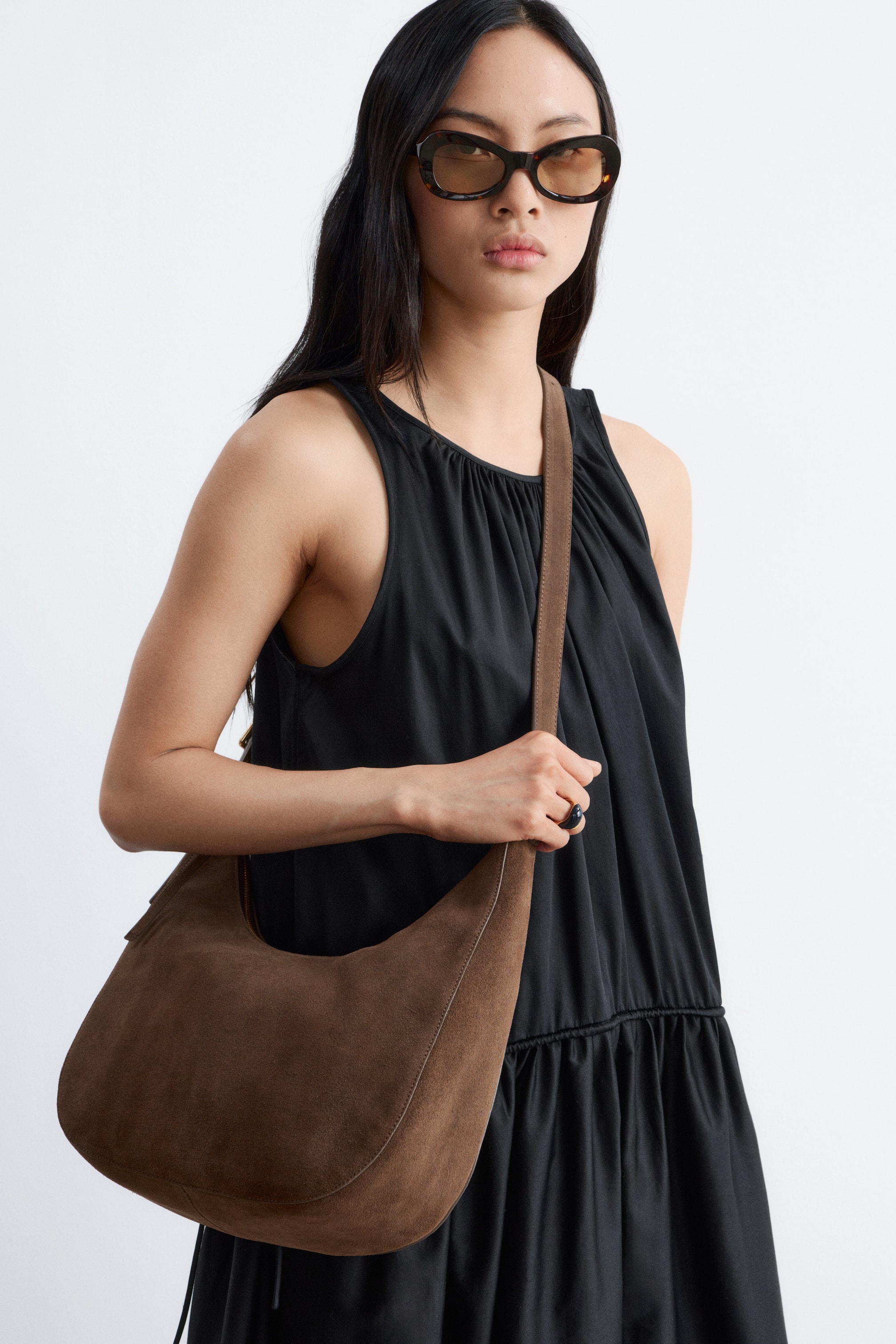 Agrandir l'image: Un sac à bandoulière en daim marron en forme de croissant est porté sur l'épaule, mettant en valeur son devant incurvé et ses coutures claires sur les bords.