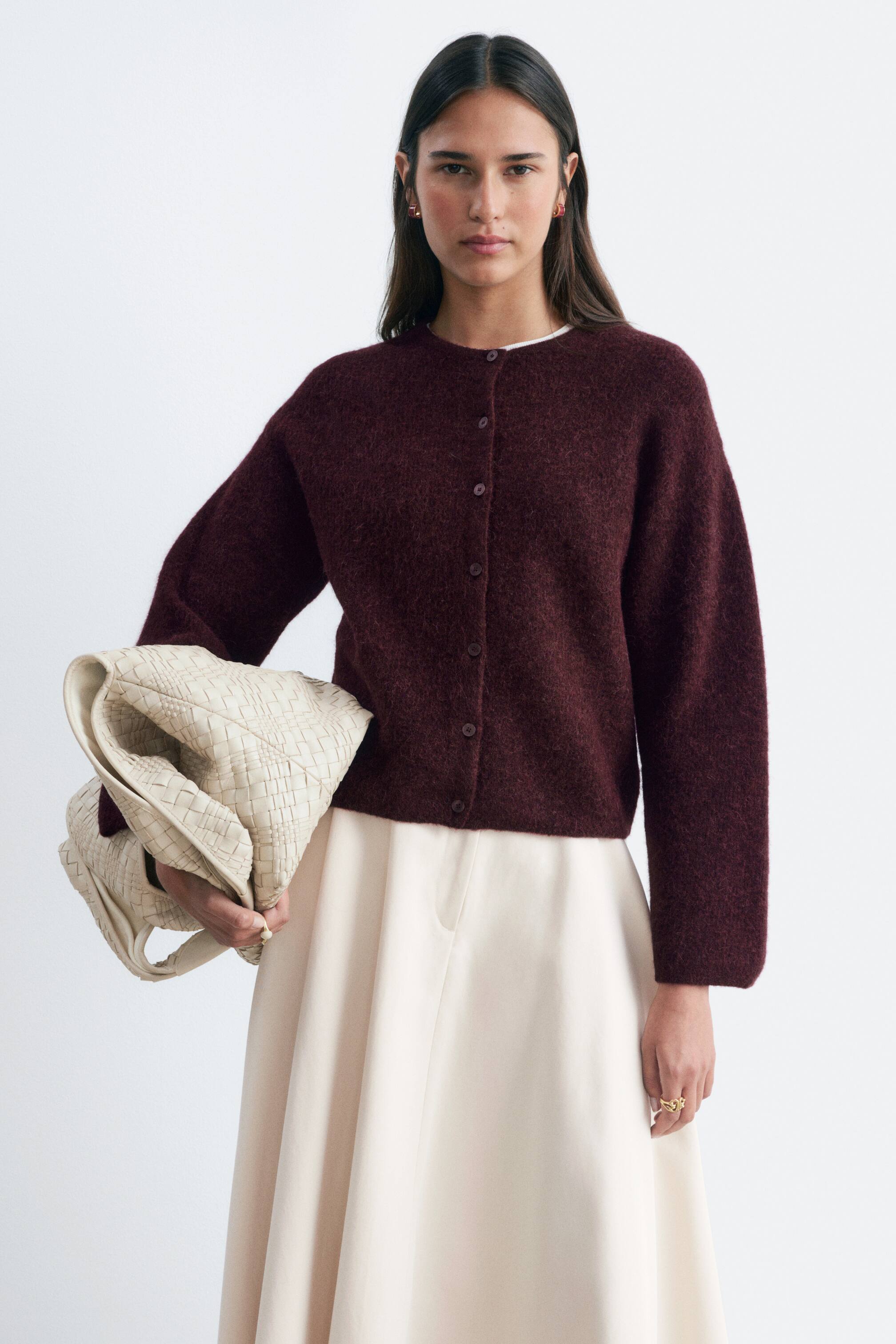 View larger image: The model is wearing a burgundy wool and alpaca blend cardigan. It boasts a round neck, long sleeves, and central front button fastenings. The model has styled the cardigan over a white top and paired it with a light-coloured skirt.