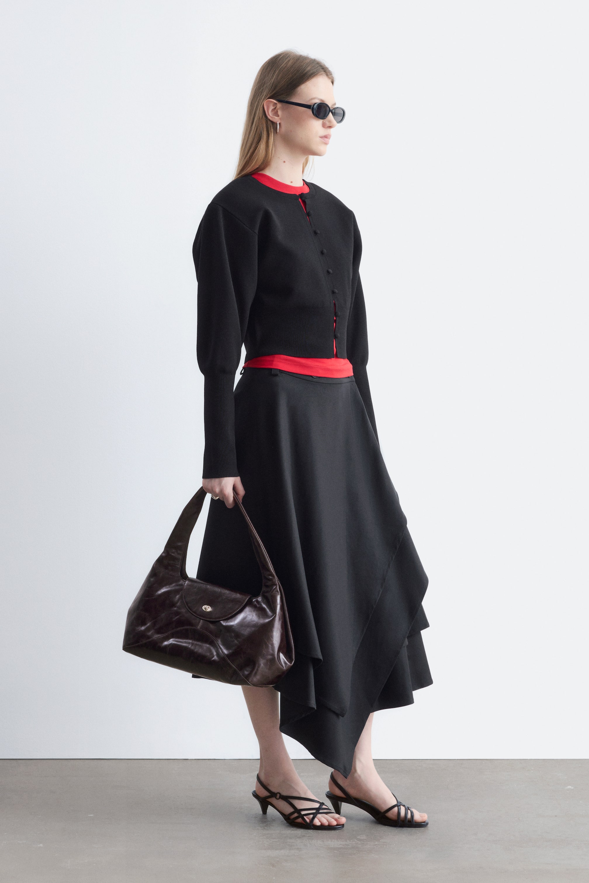 Ver imagem maior: Um cardigan preto com botões e mangas bufantes está a ser usado por cima de um top vermelho, combinado com uma saia midi assimétrica preta. A modelo também está a usar sandálias pretas de salto baixo com tiras e óculos de sol ovais, e segura uma mala de ombro em pele castanha-escura.