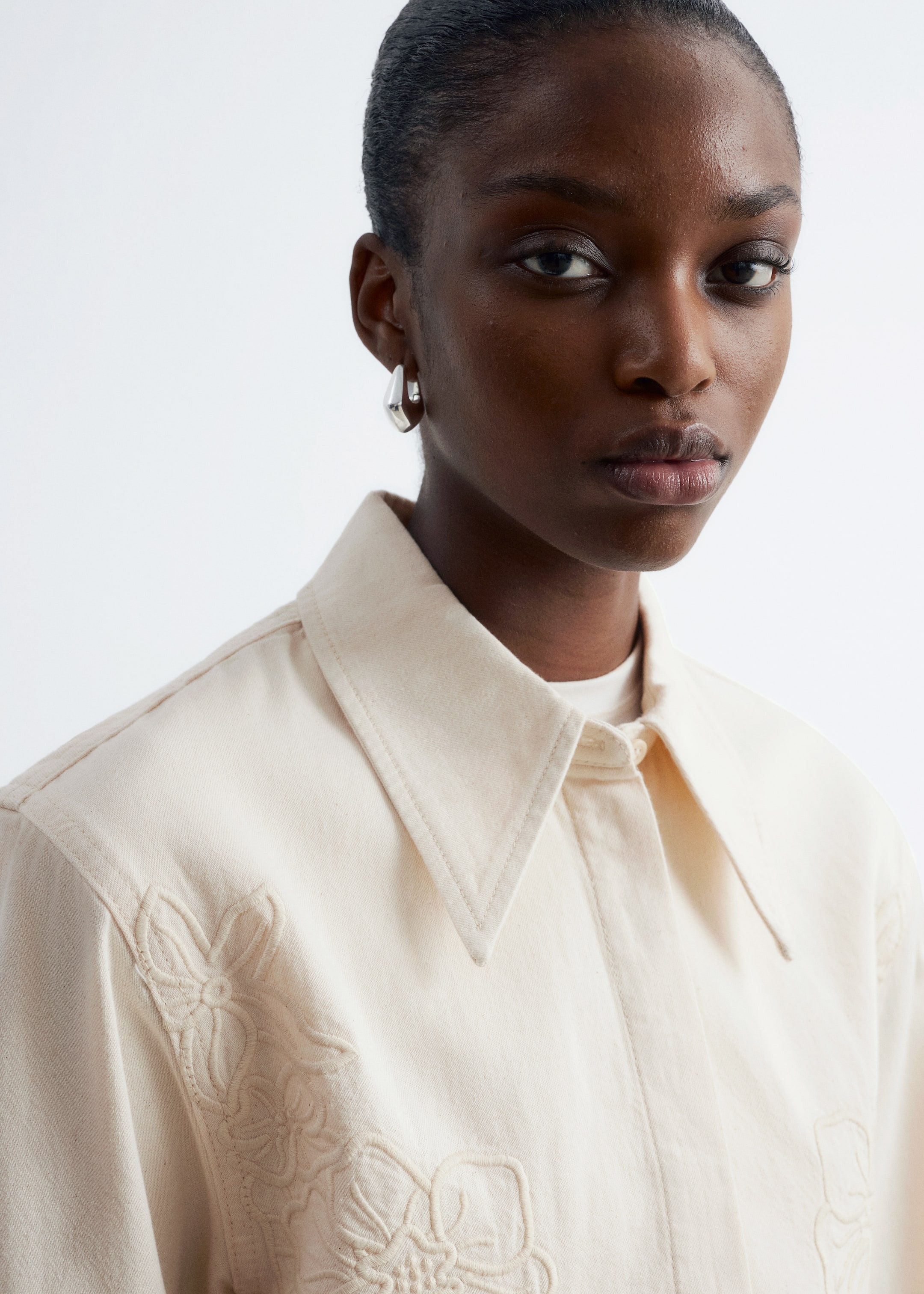 Zobrazit větší obrázek: Krémově zbarvenou džínovou košili s vyšívanými ozdobami na hrudi má na sobě modelka. Košile má klasický límec a je částečně zapnutá, odhalující bílou spodní košili pod ní.