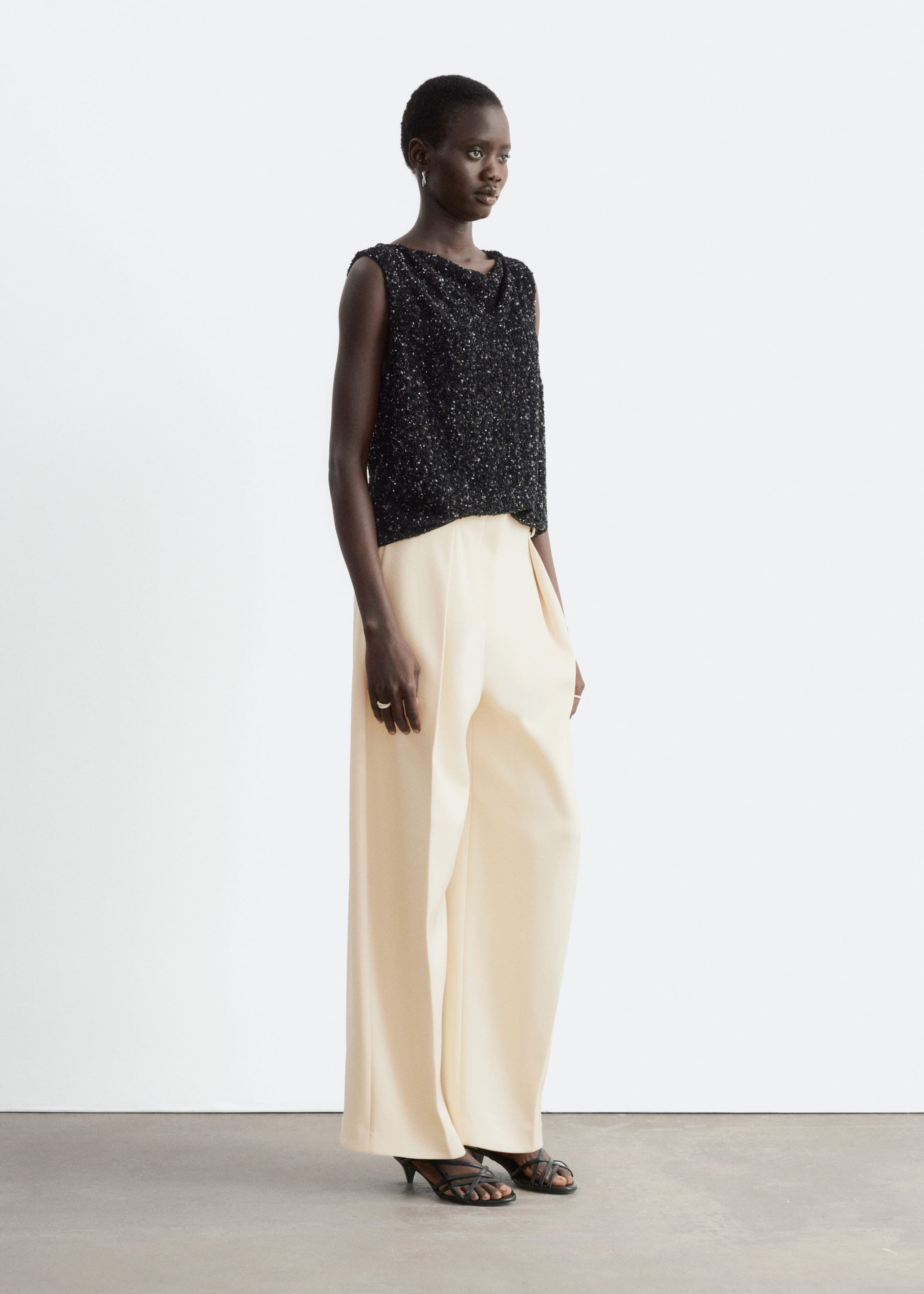 A black, sleeveless mesh top covered in shimmering sequins is being worn by the model. The top features a draped neckline and is styled with cream wide-leg trousers and black strappy sandals.