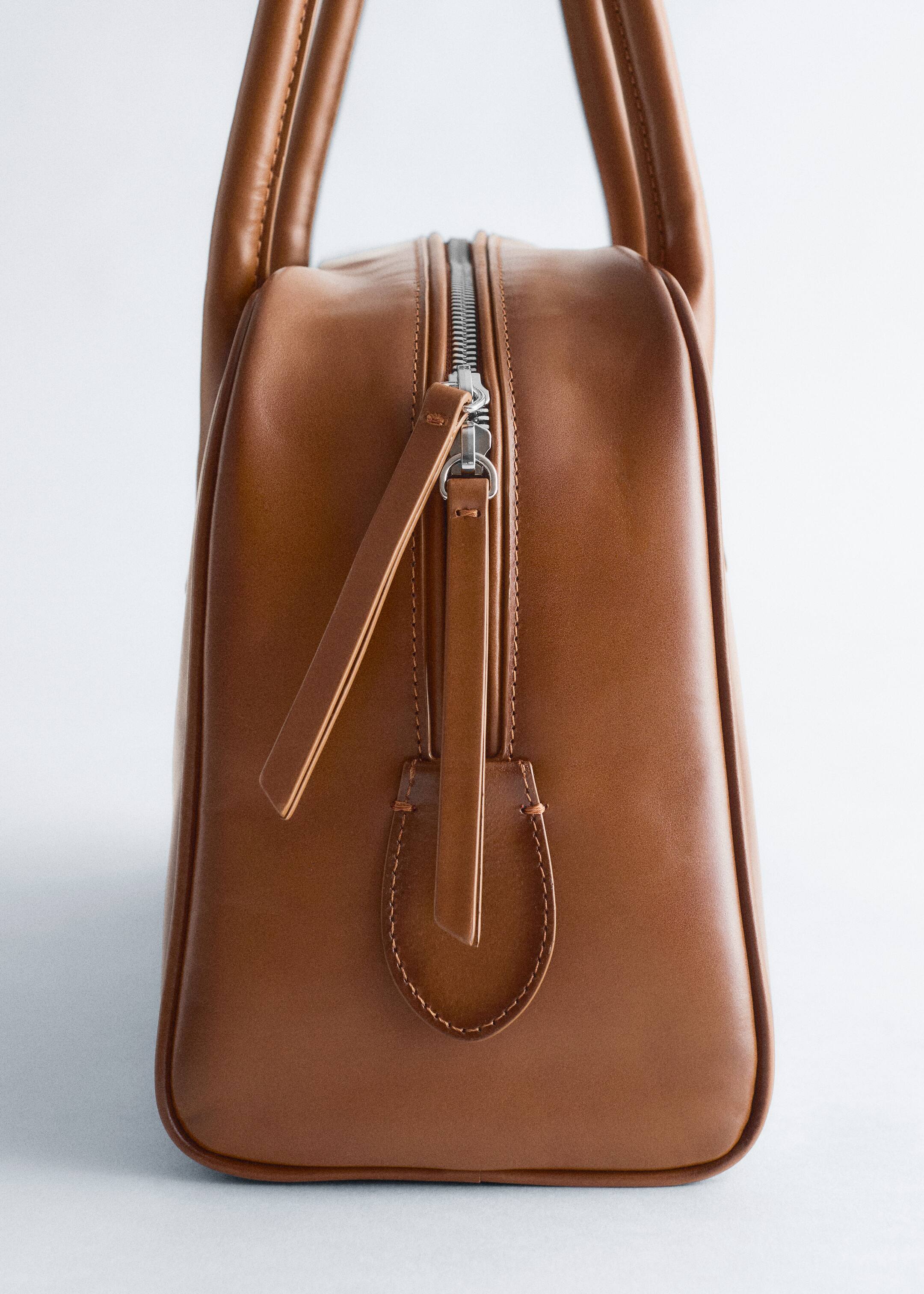 Grösseres Bild ansehen: Eine strukturierte braune Ledertasche steht nach vorne und verfügt über zwei abgerundete Griffe und einen silberfarbenen oberen Reißverschluss. Einer der beiden flachen Lederreißverschlussgriffe ist nach rechts geneigt, über einem U-förmigen Lederlasche mit sichtbaren Nähten.