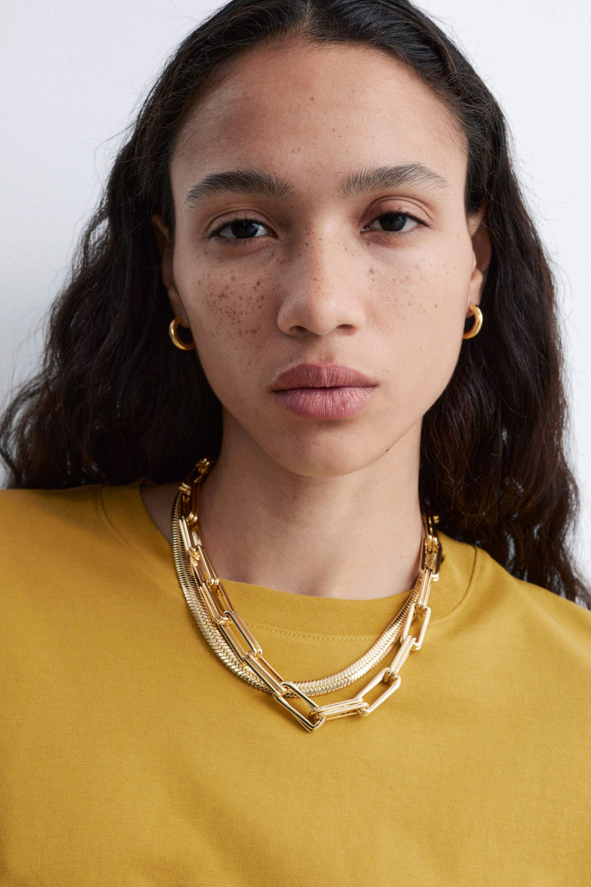 View larger image: A set of gold-toned jewellery is being presented, featuring two layered necklaces and a pair of hoop earrings. The necklaces are draping across the chest, with a chunky rectangular link chain resting over a flat snake chain, while small, thick hoop earrings are being worn on each ear.