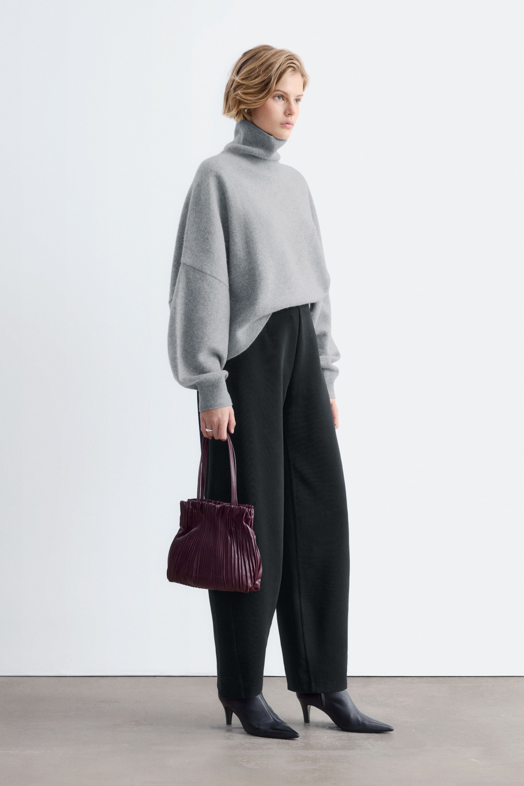 Agrandir l'image: Un pantalon noir en jersey scuba, doté d’une silhouette incurvée et de jambes larges, est porté par le mannequin. Le pantalon est associé à un pull gris oversize à col roulé, rentré à la taille, ainsi qu’à des bottes noires à talons et bout pointu.