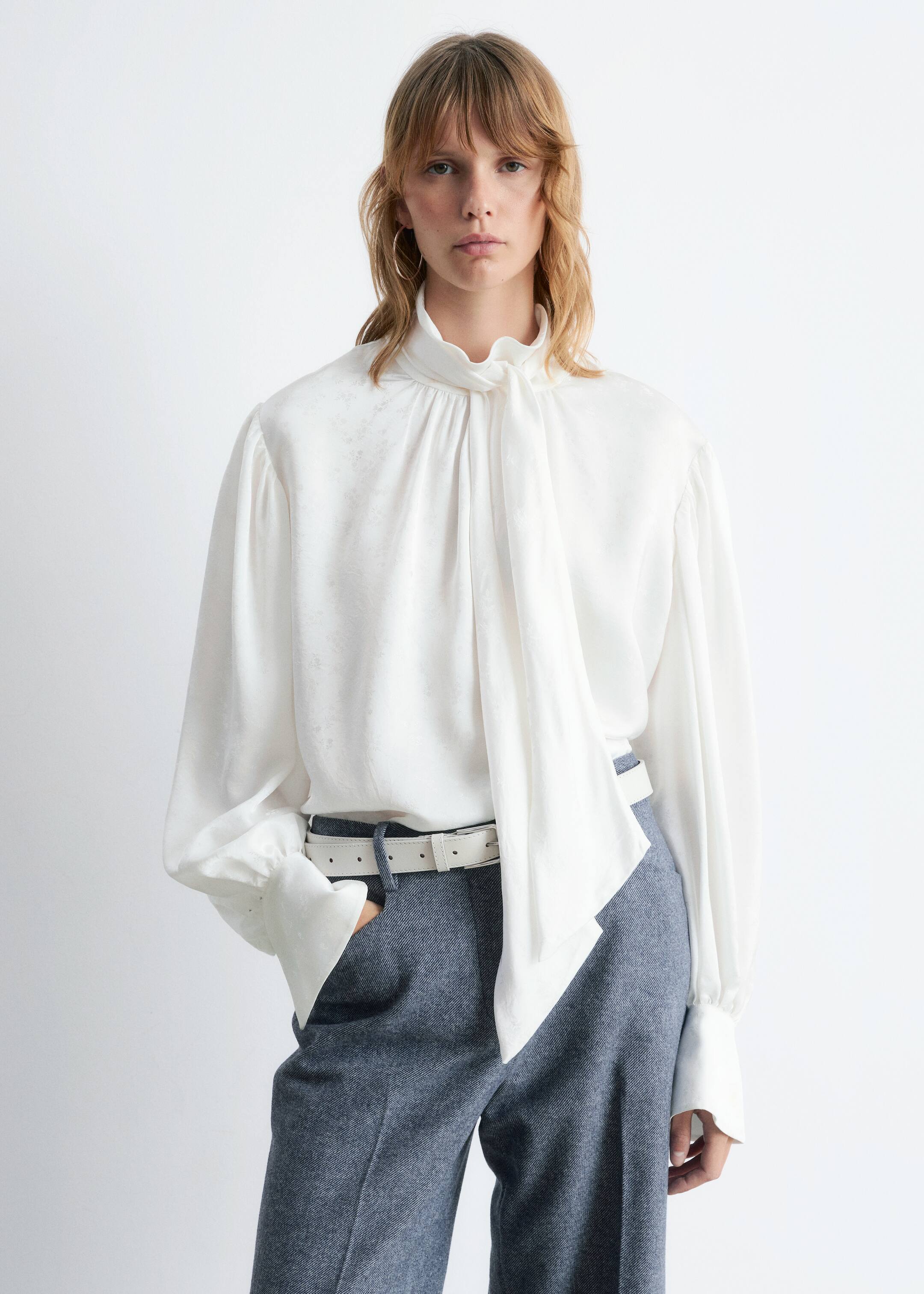 View larger image: A white jacquard blouse is featuring a high, gathered neckline with a long tie detail draping down the front. Its voluminous long sleeves are gathered at the shoulders and are finishing with wide, buttoned cuffs.