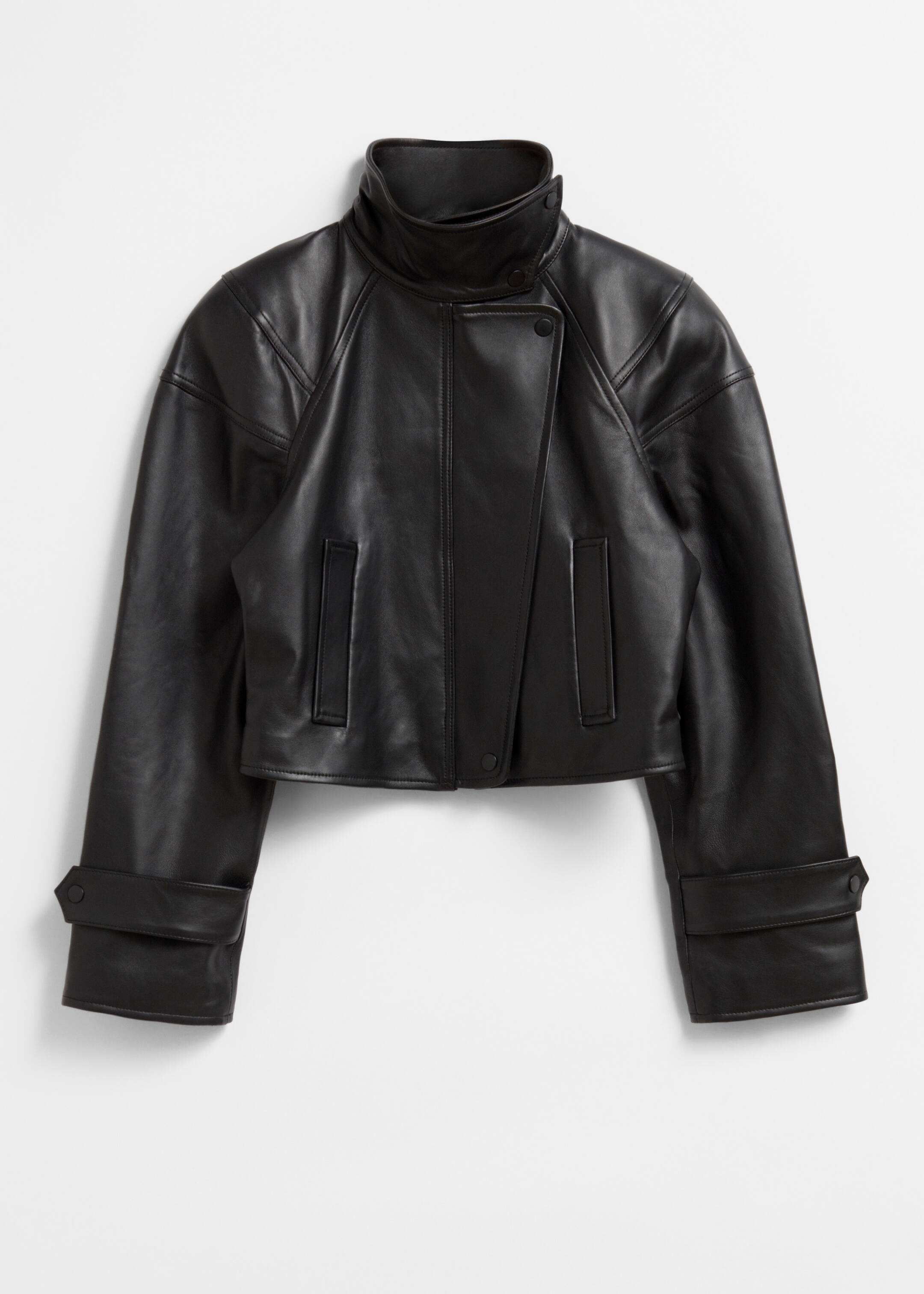 Größeres Bild ansehen: Eine schwarze, gekürzte Lederjacke wird von vorne präsentiert, mit einem glatten Finish und abgerundeten Schultern. Sie hat einen Stehkragen, einen asymmetrischen Frontverschluss mit sichtbaren Druckknöpfen, zwei rechteckige Leistentaschen und weite Ärmel mit Druckknopf-Laschen an den Manschetten.