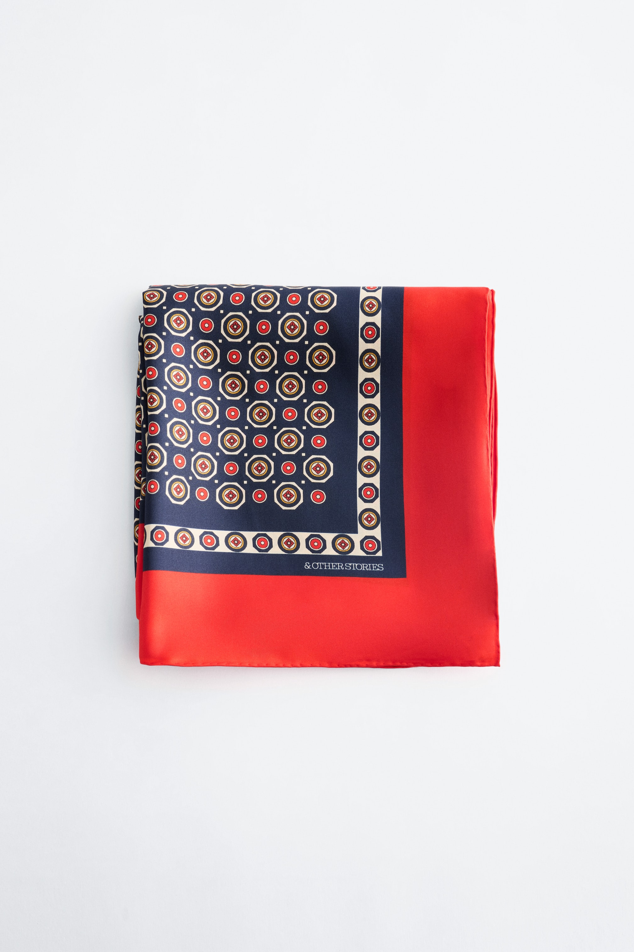 Agrandir l'image: Un foulard carré est plié, affichant une surface lustrée et présentant une section bleu marine avec un motif géométrique répétitif de cercles rouges à l'intérieur d'octogones beiges. Ce motif est bordé par une bande rouge unie le long de deux côtés, avec le texte "& OTHER STORIES" apparaissant en blanc sur la section marine.