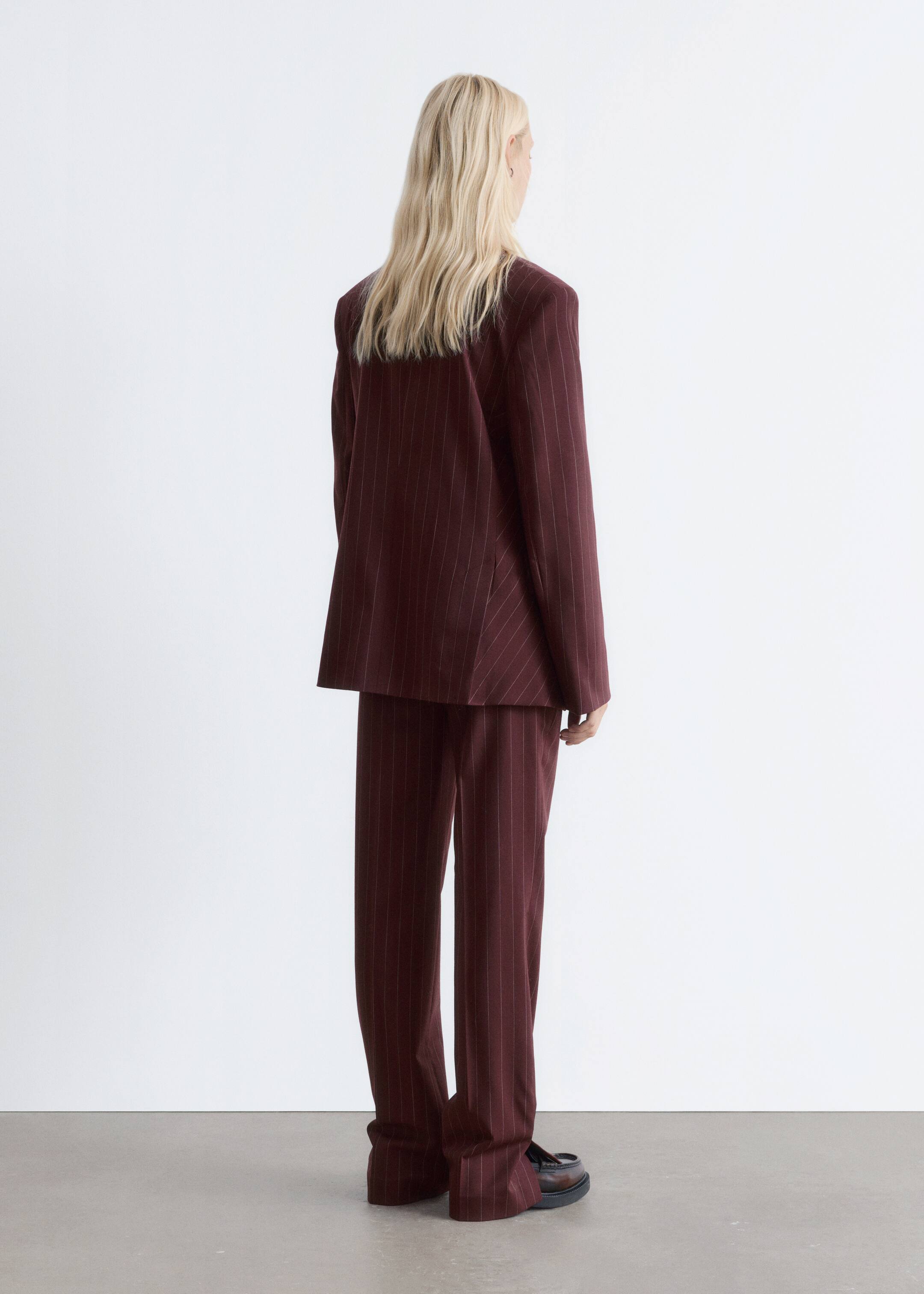 Agrandir l'image: Un costume à rayures bordeaux est vu de dos, légèrement incliné vers la droite. Le blazer surdimensionné tombe librement, et le pantalon large assorti s'accumule sur des mocassins sombres et épais.