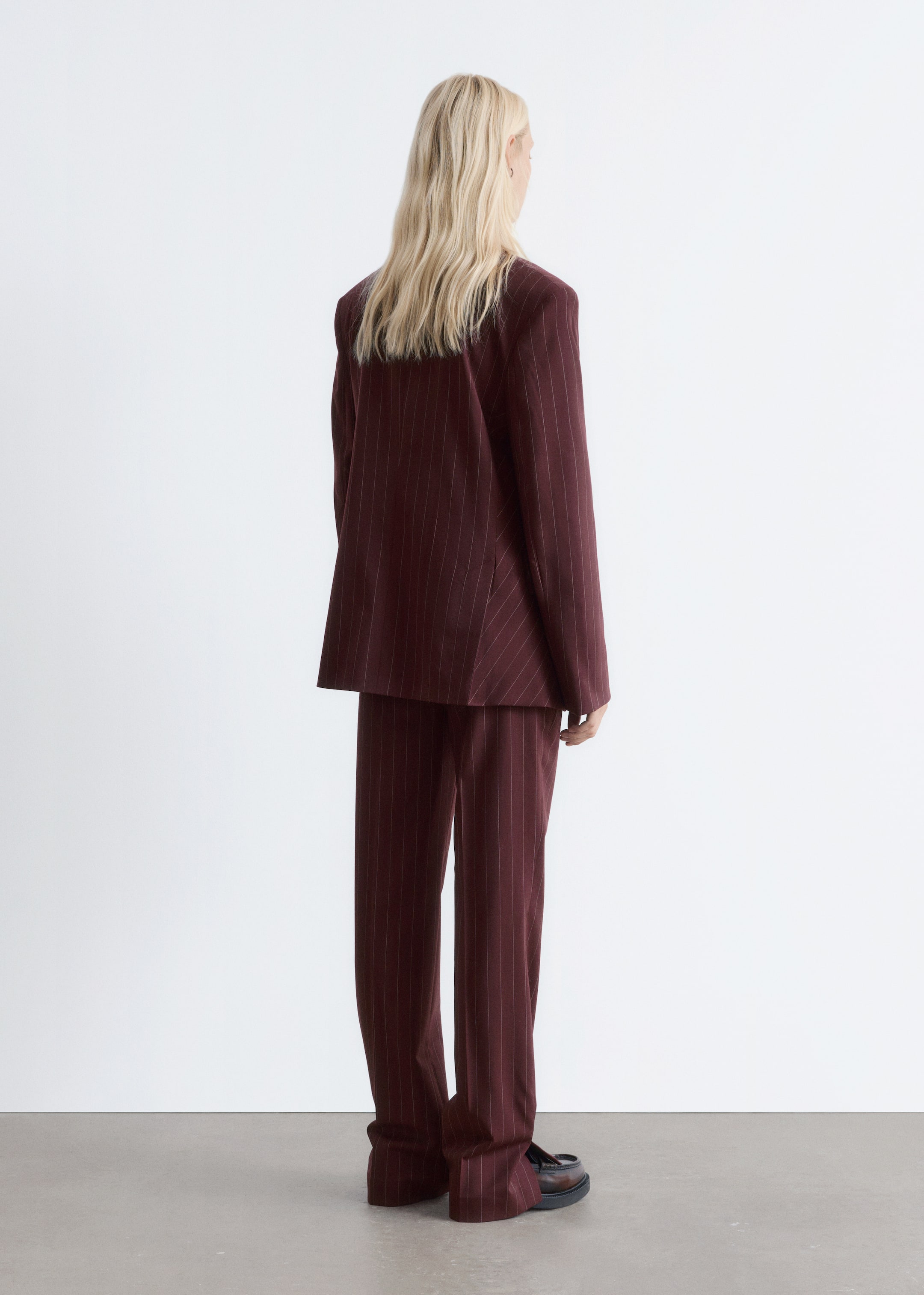 Grotere afbeelding bekijken: Een bordeauxrood krijtstreeppak wordt van achteren gezien, iets naar rechts gekanteld. De oversized blazer valt losjes en de bijpassende wijde broekspijpen vallen over donkere, grove loafers.