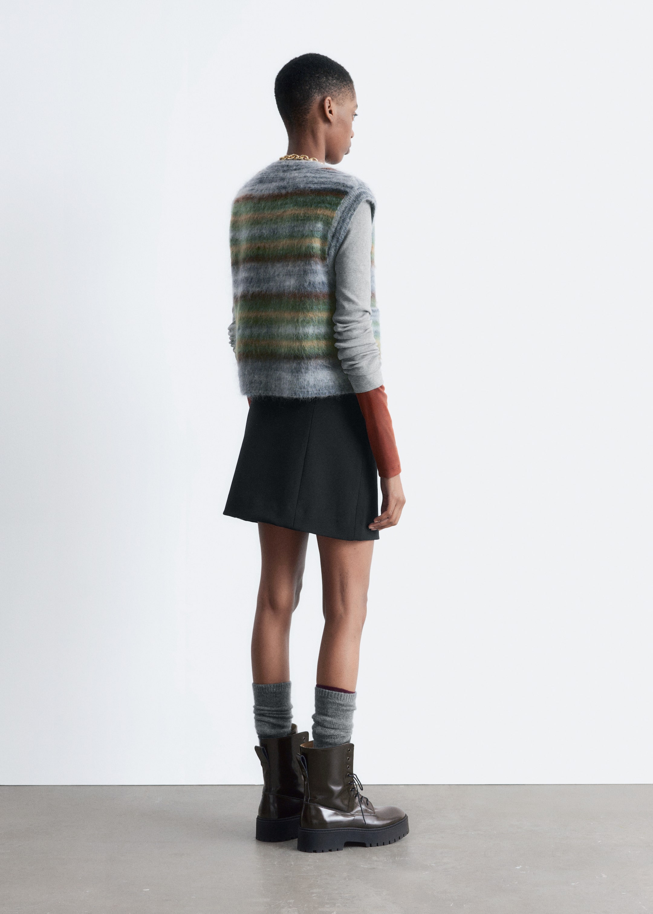 Visualizza immagine più grande: Un gilet a maglia con una finitura spazzolata e strisce orizzontali in grigio, verde e marrone è indossato dalla modella. È sovrapposto a una camicia a maniche lunghe.