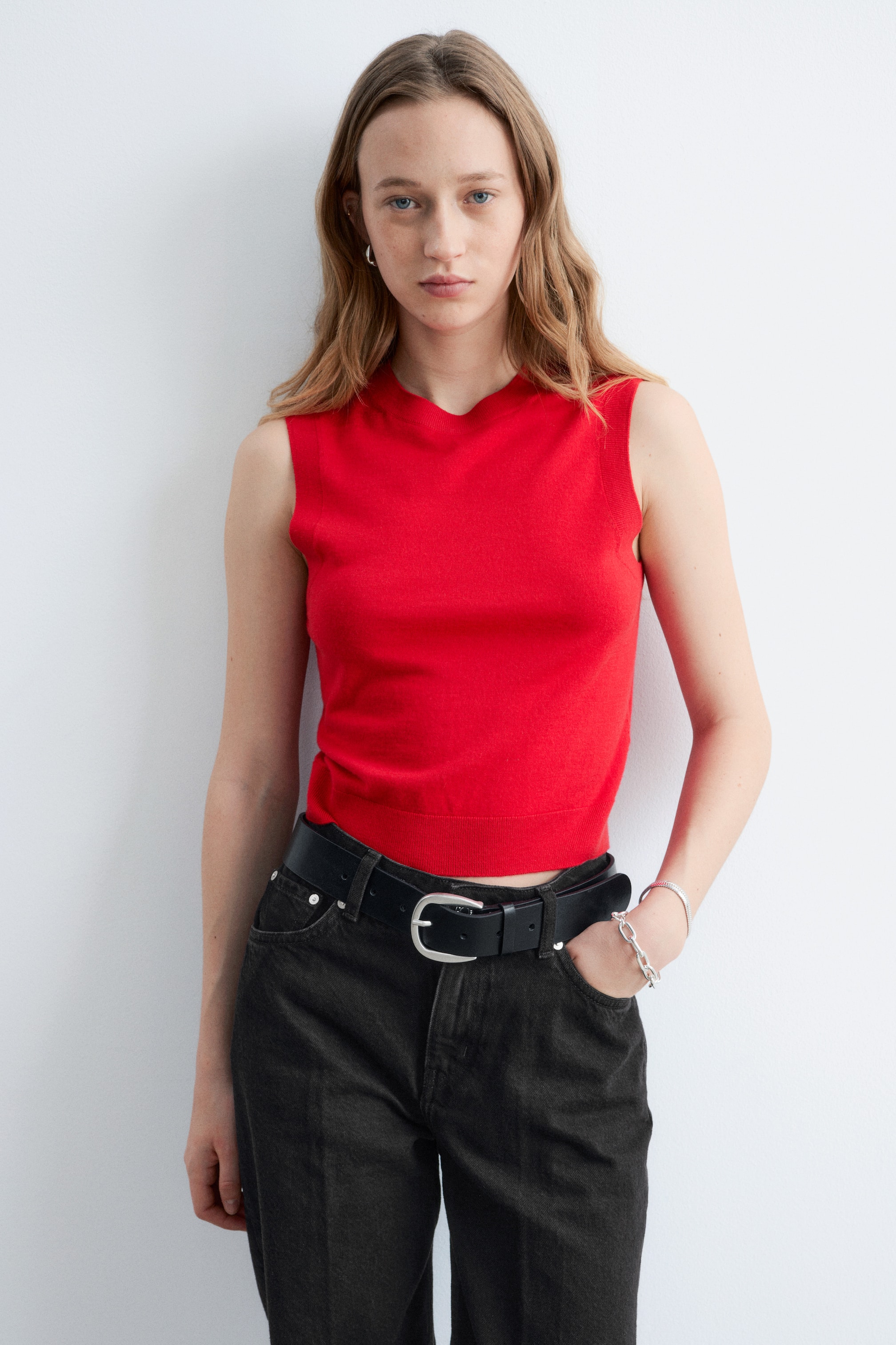 Grotere afbeelding bekijken: Een rood mouwloos gebreid topje met een gebogen V-hals en een geribde zoom, sluit nauw aan en is ingestopt in donkere jeans.