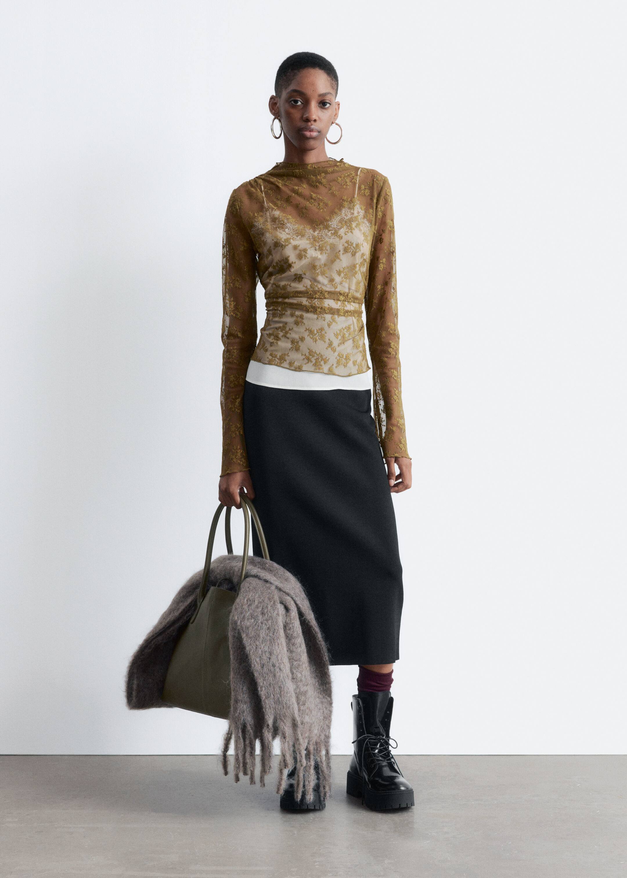Größeres Bild ansehen: Das Outfit besteht aus einem durchsichtigen langärmeligen olivgrünen Oberteil mit einem Stehkragen und einem goldenen Blumenmuster, das über einem weißen Camisole getragen wird und aus einer frontalen Perspektive präsentiert wird. Dies wird mit einem schwarzen Midi-Rock, schwarzen Schnürstiefeletten mit klobigen Sohlen und weinroten Socken sowie einer olivgrünen Tragetasche, die mit einem Fransen-besetzten, struppigen braunen Schal drapiert ist, gestylt.
