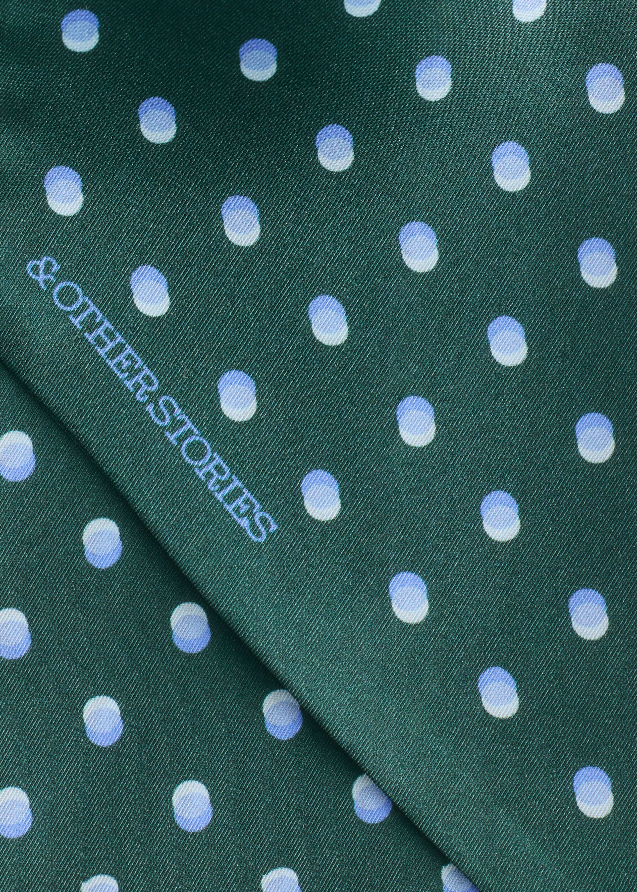 A green silk scarf features an all-over pattern of shaded light blue and white polka dots. The close-up view shows the smooth, folded fabric with the brand name "& OTHER STORIES" printed diagonally in a light blue font.