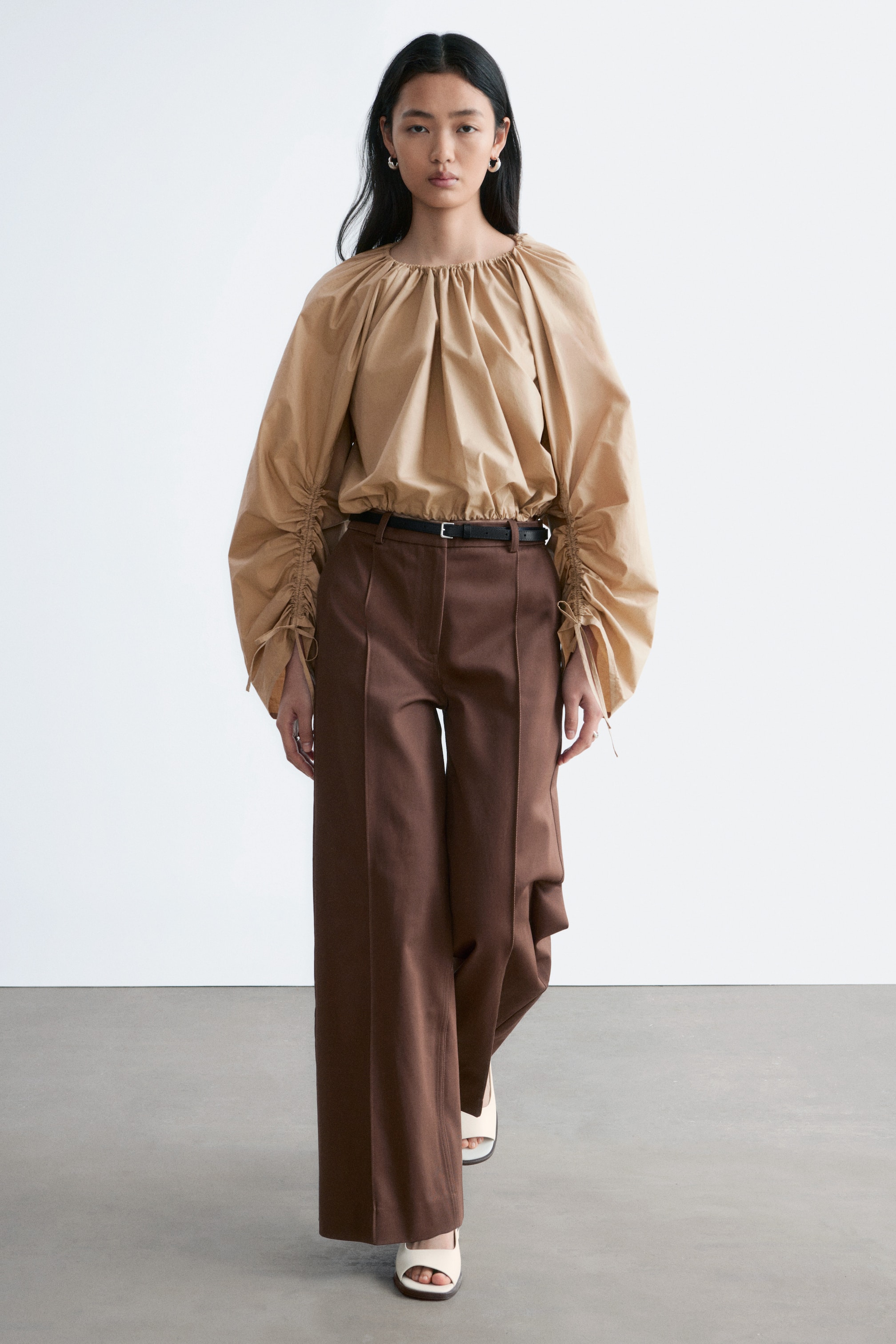 Größeres Bild anzeigen: Braune, weit geschnittene Hosen mit scharfen Bügelfalten vorne werden vom Model getragen und mit einem schwarzen Gürtel, der die Taille betont, sowie einer eingesteckten hellbraunen Bluse kombiniert.