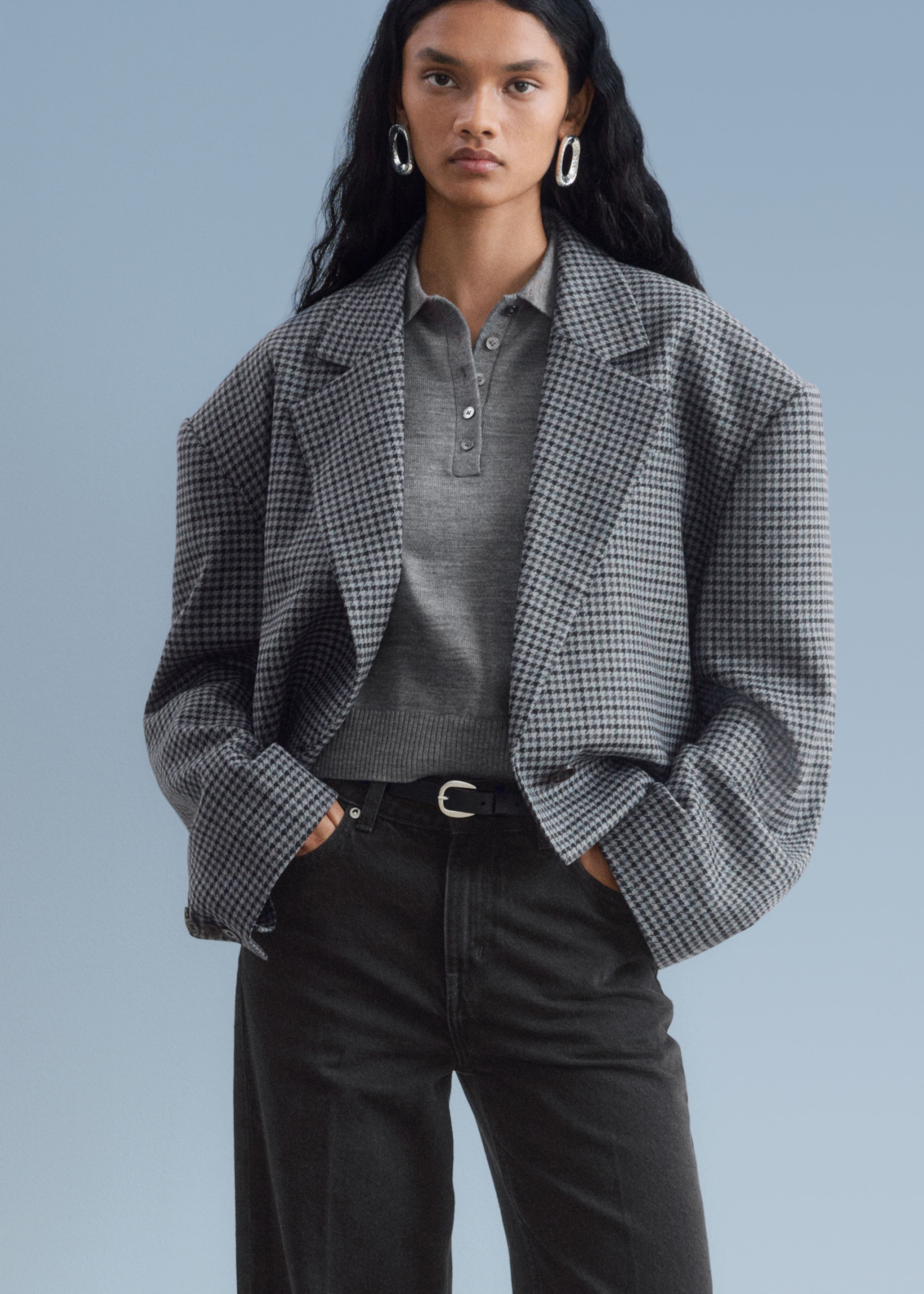 Person wearing a gray houndstooth coat over a gray shirt with buttons, paired with large silver hoop earrings.