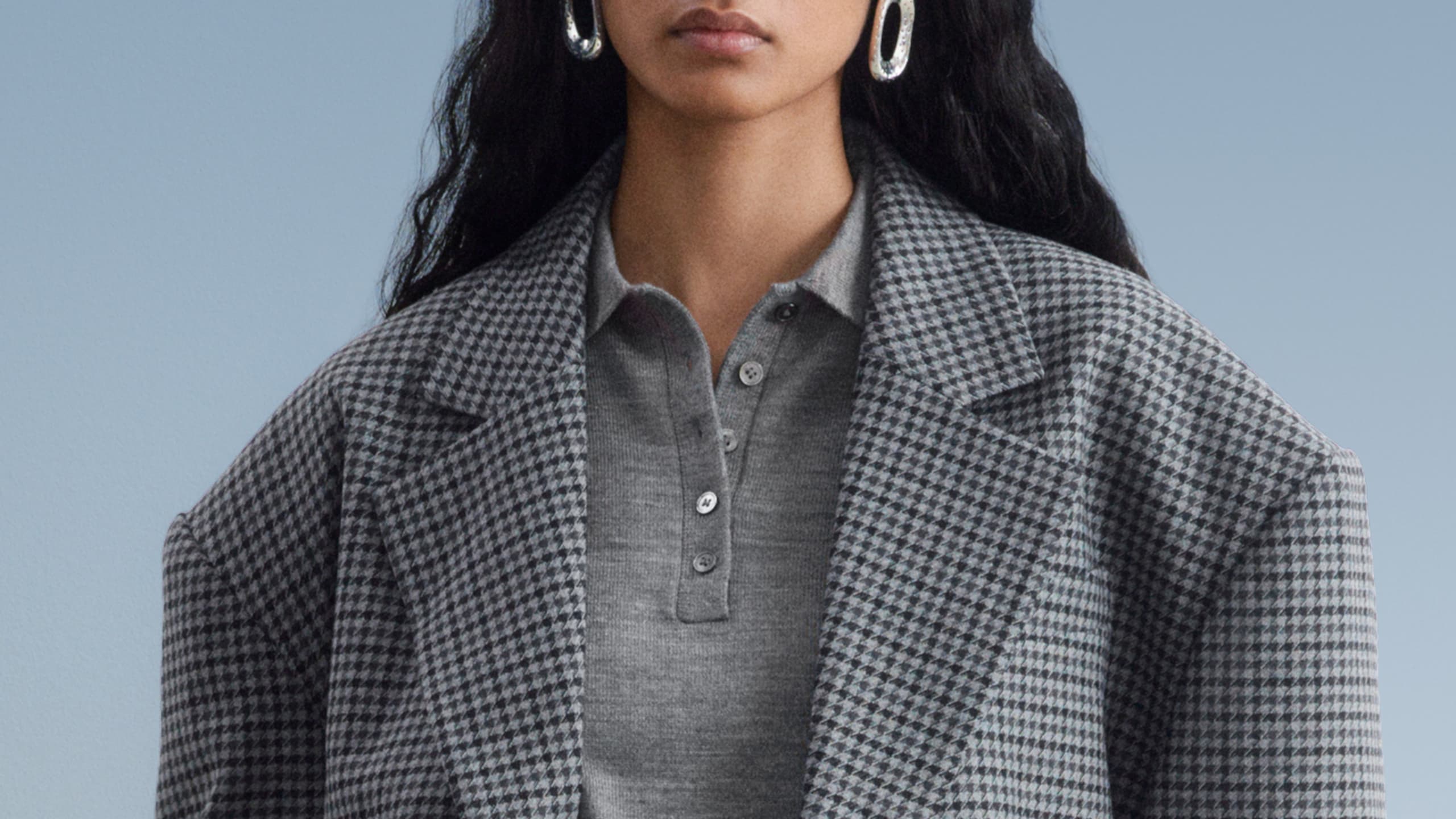 Person wearing a gray houndstooth coat over a gray shirt with buttons, paired with large silver hoop earrings.