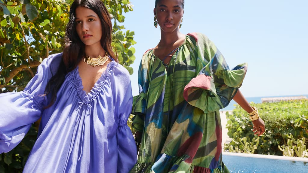 Two women in colorful, flowing dresses stand outdoors near greenery and a blue sky. One wears purple, the other a multicolored pattern.