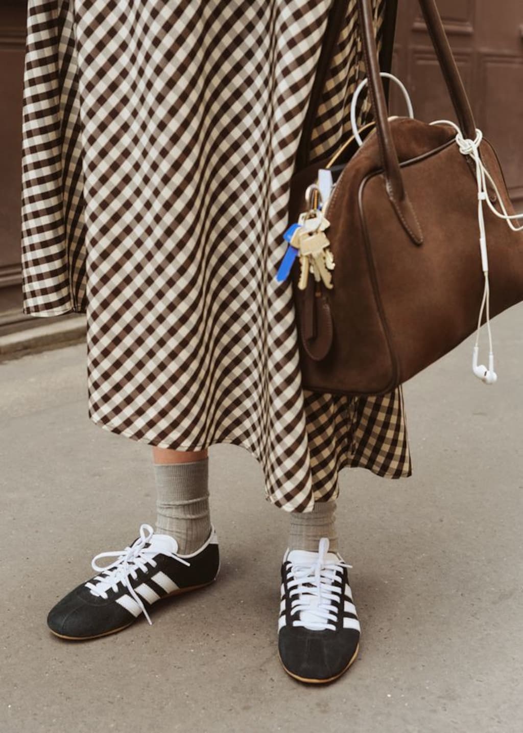 Person wearing a brown checkered skirt, black sneakers, gray socks, holding a brown bag with keys attached, standing on a sidewalk.
