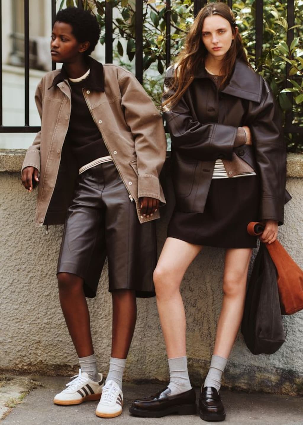 Two people in stylish outfits lean against a wall. One wears a brown jacket and shorts; the other, a dark coat and skirt. Both wear socks and sneakers.