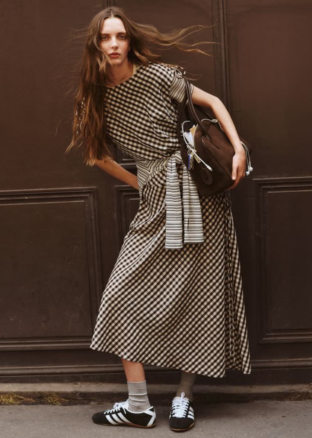 Woman in a black and white checkered dress, holding a brown bag, stands against a dark wall, wearing black sneakers and gray socks.